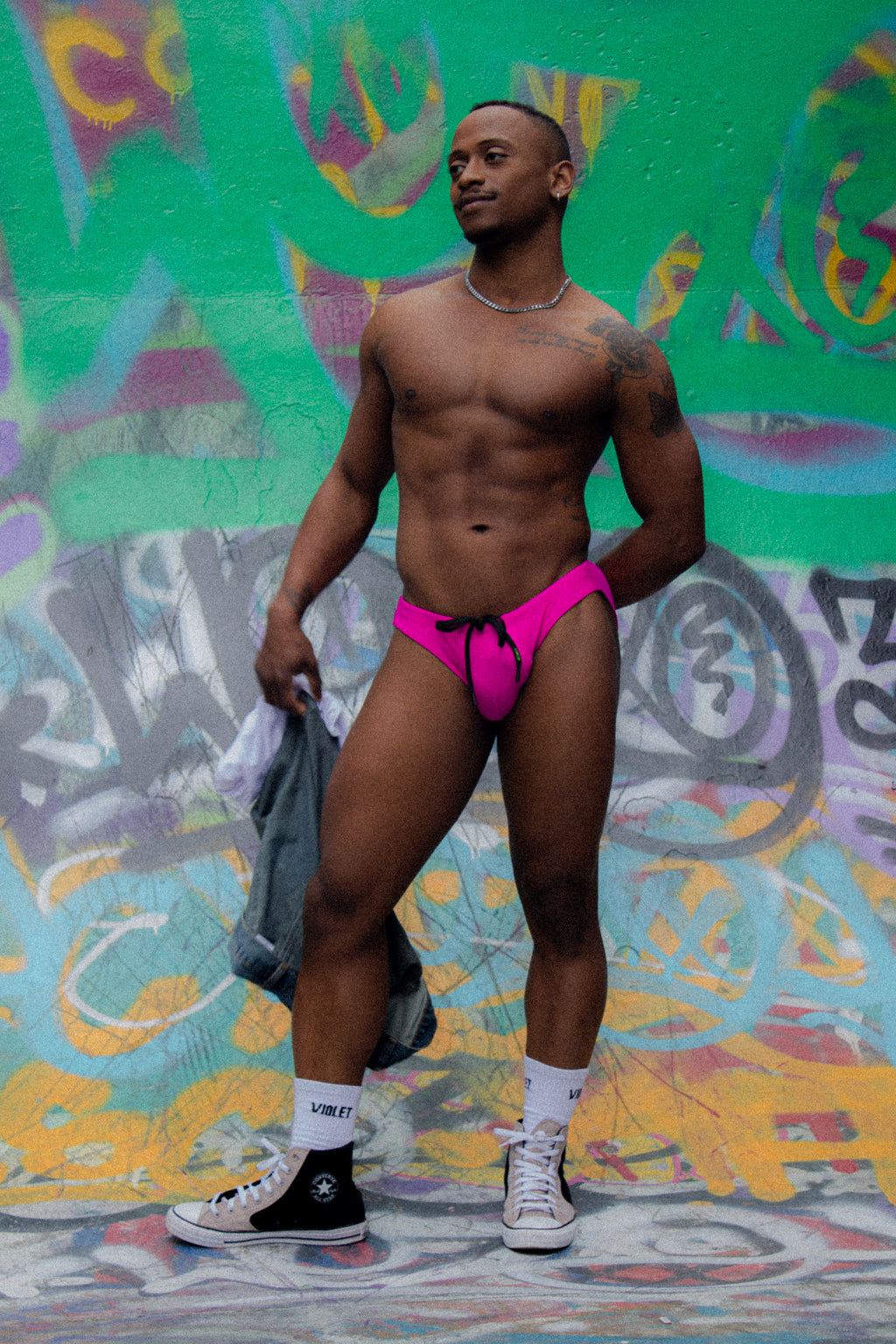 Man wearing bright pink Violet swim brief standing in front of a colorful graffiti wall.